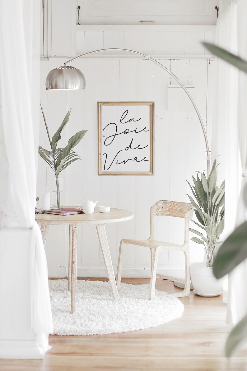 Puede incluir: Una mesa redonda blanca con una silla blanca y una alfombra blanca. Un estampado enmarcado con el texto "la joie de vivre" cuelga de la pared. Hay una planta en maceta en el suelo y una l&aacute;mpara con una pantalla plateada.