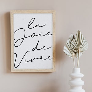 Puede incluir: Una impresi&oacute;n enmarcada con el texto "la joie de vivre" en escritura cursiva negra sobre fondo blanco. El marco es de madera clara y la impresi&oacute;n est&aacute; colgada en una pared rosa claro. Un jarr&oacute;n blanco con superficie texturizada contiene hojas de palma secas.