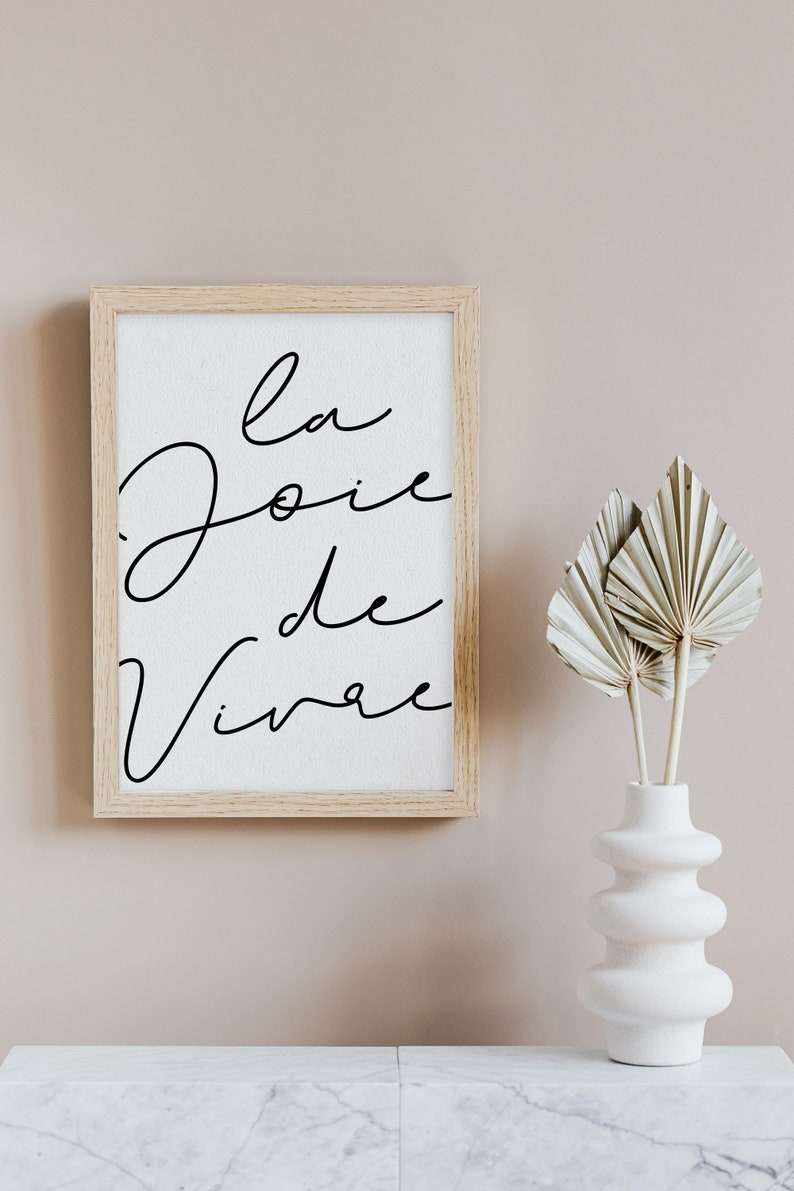Puede incluir: Una impresi&oacute;n enmarcada con el texto "la joie de vivre" en escritura cursiva negra sobre fondo blanco. El marco es de madera clara y la impresi&oacute;n est&aacute; colgada en una pared rosa claro. Un jarr&oacute;n blanco con superficie texturizada contiene hojas de palma secas.