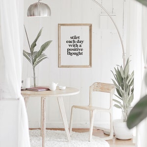 Puede incluir: Una impresi&oacute;n enmarcada en blanco con el texto "start each day with a positive thought" colgada en una pared blanca en una sala de estar minimalista. La habitaci&oacute;n cuenta con una mesa blanca con una silla blanca, una alfombra blanca y una planta en maceta.