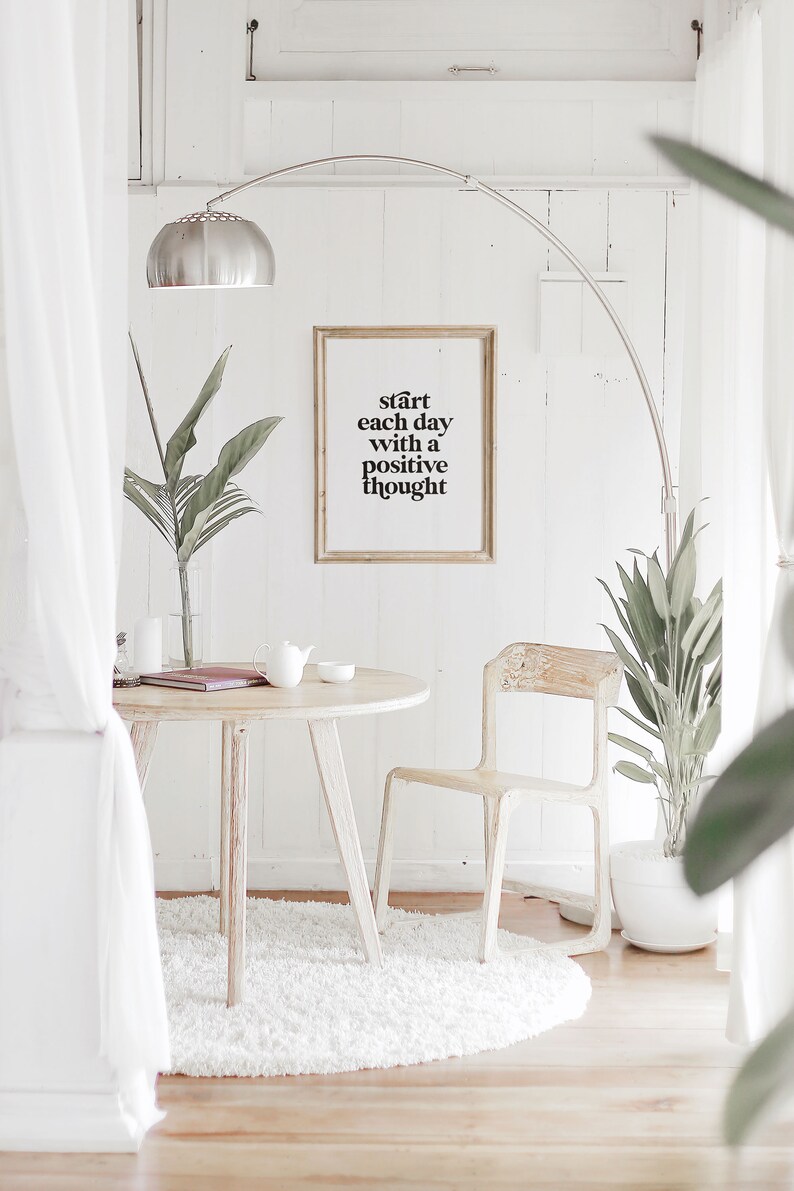 Puede incluir: Una impresi&oacute;n enmarcada en blanco con el texto "start each day with a positive thought" colgada en una pared blanca en una sala de estar minimalista. La habitaci&oacute;n cuenta con una mesa blanca con una silla blanca, una alfombra blanca y una planta en maceta.