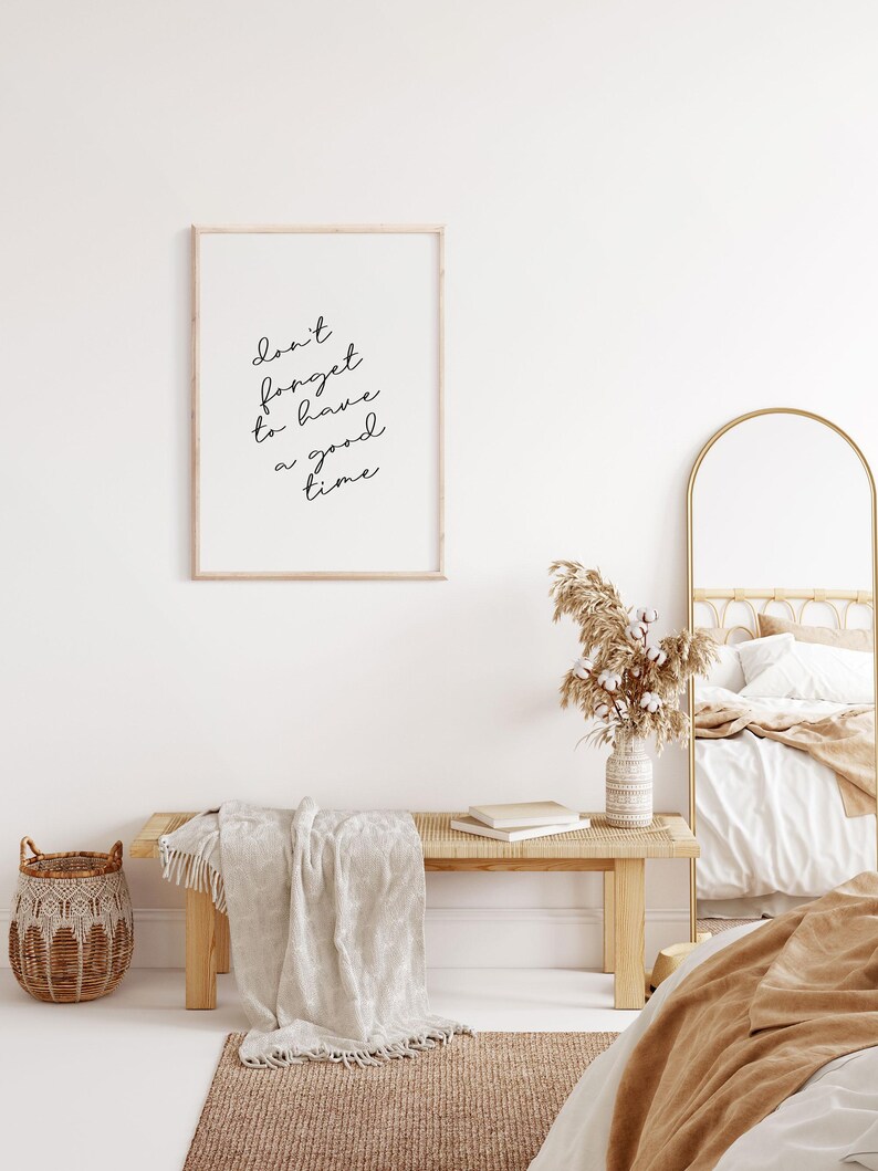 May include: A framed print with the text "Don't forget to have a good time" in a minimalist bedroom setting. The print is hanging on a white wall above a wooden bench with a white throw blanket. A vase with dried pampas grass is on the bench.