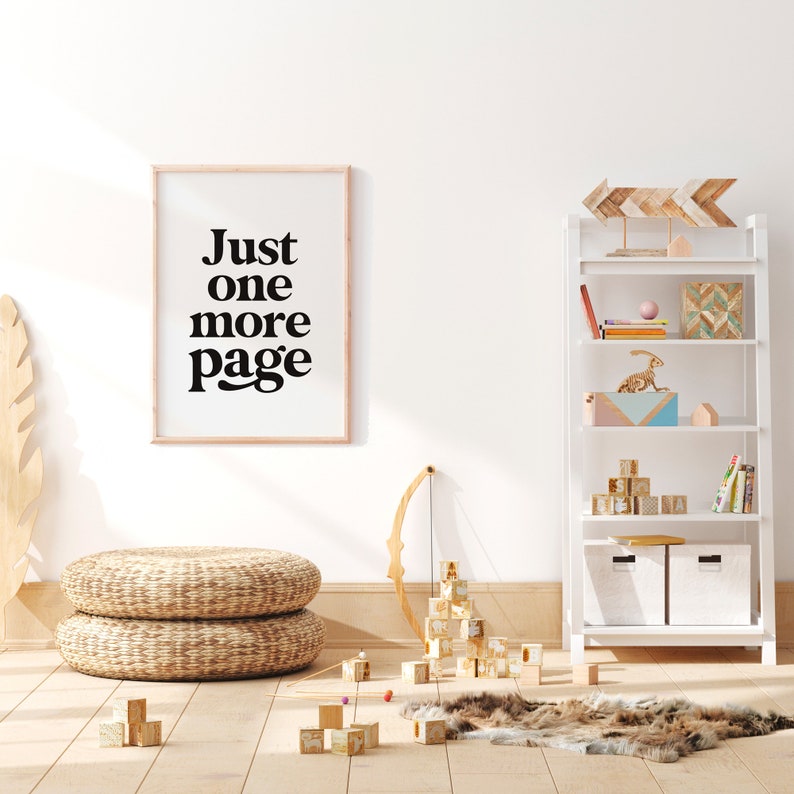 May include: A white framed print with black text that reads "Just one more page" hanging on a white wall. A white bookshelf with wooden shelves is on the right side of the image. The bookshelf is decorated with wooden arrows, a small wooden house, a colorful geometric box, a pink and blue box, and a wooden dinosaur. There are wooden blocks stacked on the shelves and a few wooden blocks on the floor. A woven wicker ottoman is on the left side of the image. A wooden bow and arrow is leaning against the wall.