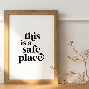 May include: A framed print with the text "this is a safe place" in black bold letters on a white background. The frame is light brown wood.