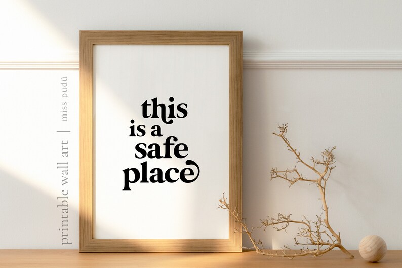 May include: A framed print with the text "this is a safe place" in black bold letters on a white background. The frame is light brown wood.