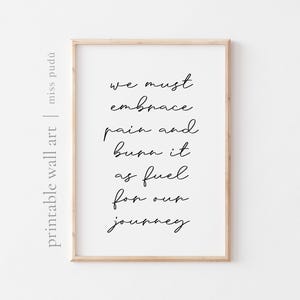 Puede incluir: Una impresión enmarcada con un marco de madera clara. La impresión presenta texto negro en cursiva sobre un fondo blanco que dice: "debemos abrazar el dolor y quemarlo como combustible para nuestro viaje".