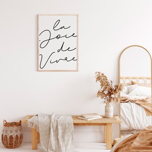 Puede incluir: Una impresi&oacute;n enmarcada con el texto "la joie de vivre" en escritura negra sobre fondo blanco. La impresi&oacute;n est&aacute; colgada en una pared blanca sobre un banco de madera con una manta blanca drapeada sobre &eacute;l. Un jarr&oacute;n con hierba de la pampa seca est&aacute; en el banco. Un espejo de arco de metal dorado est&aacute; en el fondo.