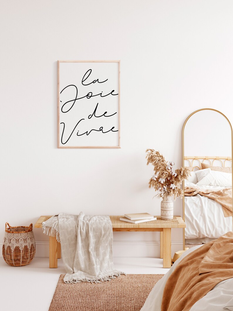 Puede incluir: Una impresi&oacute;n enmarcada con el texto "la joie de vivre" en escritura negra sobre fondo blanco. La impresi&oacute;n est&aacute; colgada en una pared blanca sobre un banco de madera con una manta blanca drapeada sobre &eacute;l. Un jarr&oacute;n con hierba de la pampa seca est&aacute; en el banco. Un espejo de arco de metal dorado est&aacute; en el fondo.