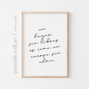 May include: A framed print with the text "un hogar sin libros es como un cuerpo sin alma" in black script on a white background. The frame is a light wood color. The words "printable wall art" and "miss pudú" are printed on the left side.
