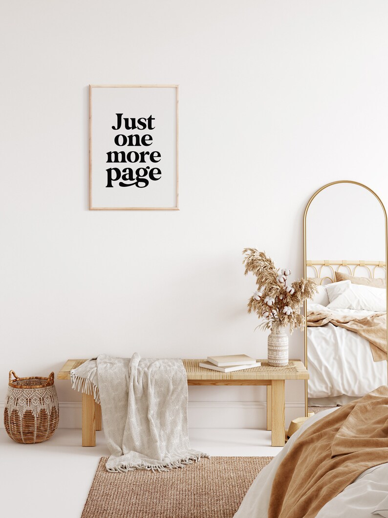 May include: A white wall with a framed print that reads "Just one more page". A wooden bench with a white throw blanket sits in front of the wall. A wicker basket sits on the floor next to the bench. A gold arched mirror and a bed with a white and brown blanket are in the background.