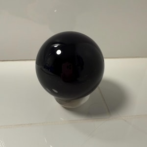May include: A shiny black sphere, possibly a decorative item or a paperweight, rests on a small, white base. The sphere reflects light, creating bright white highlights. The object is positioned on a white tiled surface.