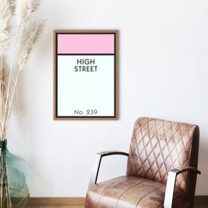 May include: A framed print featuring a pink and light blue design with the words "HIGH STREET" in black text. Below the text is the number "No. 239". The print is in a wooden frame and hangs on a white wall.