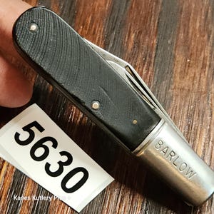 May include: A closed folding pocket knife with a black textured handle and a silver metal bolster engraved with the word BARLOW. A white tag with the black printed number 5630 and the text Kases Kutlery Plus is positioned next to the knife.