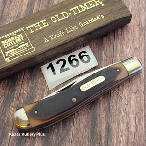 May include: A pocket knife with a brown and black handle and a silver blade. The knife is labeled "OLD TIMER" and is next to a wooden box with the text "THE OLD-TIMER" and "A Knife Like Grandad's". A sticker with the number "1266" is also present.