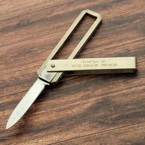May include: A vintage folding knife with a silver handle and blade. The handle has a rectangular loop design. The knife's handle has the text "GENERAL MOTORS CLUB OF ATLANTA". The knife is partially open, revealing the blade.