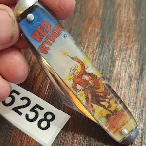 May include: A folding pocket knife with a handle featuring a graphic of a cowboy on a horse holding a torch, with the text RED RYDER printed in red. The knife has silver-colored metal bolsters and rivets, and a white tag with the number 5258.