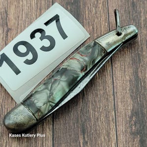 May include: A vintage pocket knife with a mother-of-pearl handle and silver-toned accents. The knife is partially open, revealing the blade. The number "1937" is visible on a white label. The text "Kases Kutlery Plus" is also visible.
