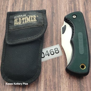 May include: A black nylon sheath and a folding knife with a green handle and a silver blade. The sheath has the text "SCHRADE OLD TIMER U.S.A." The knife is partially open, revealing the blade. A white tag with the number "0468" is also visible.