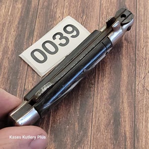 May include: A closed black pocket knife with a silver bolster and rivets. A white tag with the number "0039" is on top of the knife. The knife is on a wooden surface. The text "Kases Kutlery Plus" is at the bottom left.