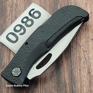 May include: A black folding knife with a textured handle and a silver blade. The knife has a small circular emblem and a partially visible white label with the number "0986". The text "Kases Kutlery Plus" is at the bottom.