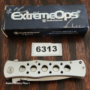 May include: A silver folding knife with a textured handle featuring five circular cutouts, positioned below a rectangular box labeled ExtremeOps Smith & Wesson and a white sticker with the number 6313 printed in black text.
