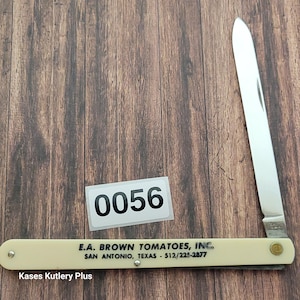 May include: A vintage folding knife with a cream-colored handle and a stainless steel blade. The handle has the text "E.A. BROWN TOMATOES, INC. SAN ANTONIO, TEXAS - 512/225-2877". A sticker with the number "0056" is also present. The knife is open, displaying the blade.