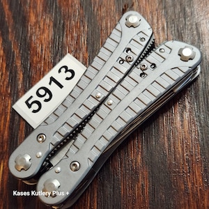 May include: A grey multi-tool with a textured handle and visible screws, featuring the text 5913 on a white rectangular tag and the text Kases Kutlery Plus + printed at the bottom. The tool is in a closed position with a metallic finish.