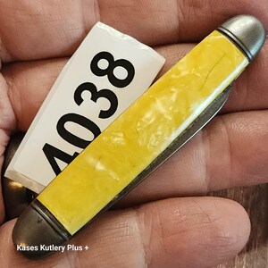 May include: A closed pocket knife featuring a bright yellow, marbled handle with metallic silver-toned bolsters at both ends. A white rectangular tag with the black printed number 4038 is positioned alongside the knife handle.