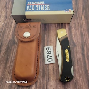 May include: A Schrade Old Timer folding knife with a black handle and a gold-colored bolster, next to a brown leather sheath. The knife is partially open, revealing the blade. The box is visible in the background.