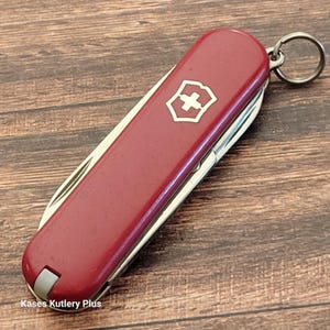 May include: A red Swiss Army knife with a silver keyring. The knife features a white cross emblem on a red shield. The multi-tool is closed, with several tools visible along the side. The text "Kases Kutlery Plus" is visible at the bottom.