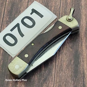 May include: A folding pocket knife with a dark wood handle and a silver blade. The knife has a metal bolster and a loop for attaching to a keychain. The number "0701" is printed on a white label. The text "Kases Kutlery Plus" is also visible.
