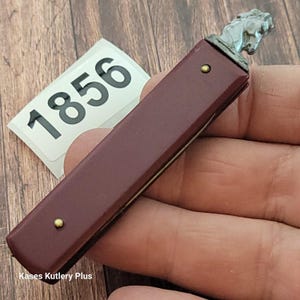 May include: A burgundy pocket knife with a horse head design on the handle. The knife has brass rivets and a silver-colored horse head. The number "1856" is visible on a white label. The text "Kases Kutlery Plus" is also visible.