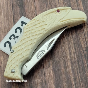 May include: A cream-colored pocket knife with an eagle head design. The knife blade is silver, and the handle is carved to resemble an eagle's head with a red eye. The text "200" is visible on the blade. The text "Kases Kutlery Plus" is visible at the bottom.