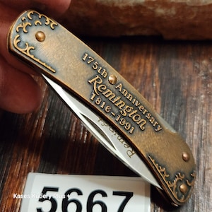 May include: A bronze-toned folding pocket knife featuring an engraved 175th Anniversary Remington 1816-1991 commemorative design on the handle. The handle includes decorative scrollwork at the ends and two visible rivets. A sharp steel blade is partially visible.