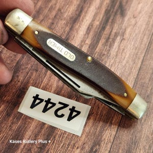 May include: A closed Old Timer pocket knife with a brown handle, silver-toned metal bolsters, and brass pins. The handle features a textured dark brown inlay and a white oval shield with the text OLD TIMER. A white tag displays the number 4244.