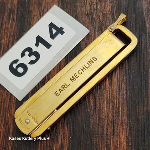May include: A gold-toned metal money clip featuring the engraved name EARL MECHLING and the text EXECUTIVE MADE IN U.S.A. The clip is positioned next to a white rectangular tag with the black printed number 6314.