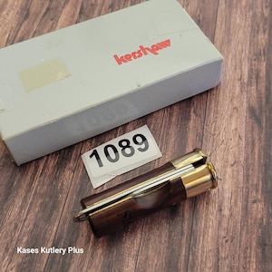 May include: A folding pocket knife with a dark wood handle and a brass bolster, next to a gray box with the red "Kershaw" logo. A white sticker with the number "1089" is also present. The knife is partially open, revealing the blade.