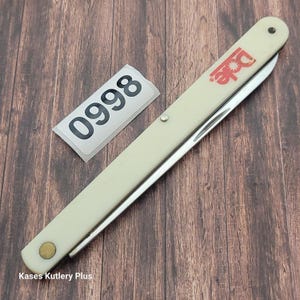 May include: A vintage fruit knife with a cream-colored handle and a stainless steel blade. The knife features the Dole logo in red. A white sticker with the number 0998 is next to the knife.
