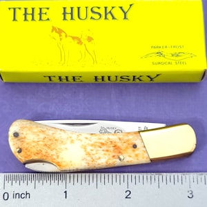 May include: A small pocket knife with a bone handle and a brass bolster. The blade is closed and the knife is about 3 inches long. The knife is on a ruler with the text "THE HUSKY" on the box.