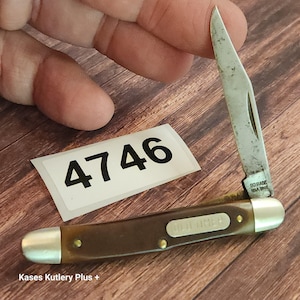 May include: A folding pocket knife with a brown handle, silver-colored metal bolsters, and a single open blade. The blade is stamped with the text SCHRADE USA 194T, and the handle features an Old Timer shield. A white sticker with the number 4746 is present.