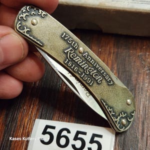 May include: A folding pocket knife with a brass-toned metal handle featuring decorative scrollwork at the ends. The handle is engraved with the text 175th Anniversary Remington 1816-1991. A partially visible silver-colored metal blade extends from the handle.