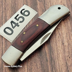 May include: A folding pocket knife with a dark wood handle and a stainless steel blade. The handle is secured with brass rivets. A white label with the number "0456" is visible.