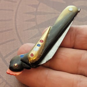 May include: A small, antique folding knife with a dark handle and a gold-colored bolster. The bolster is decorated with red, white, and blue dots. The blade is silver and partially open, revealing the sharp edge. The knife has a unique, stylized design.