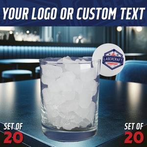 May include: Set of 20 clear ice cubes in a glass on a dark table. The ice cubes are in a bar setting. A white round coaster with a blue and red logo that says "Lasercraft Company" is on the table.