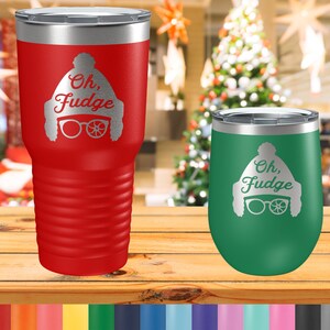 May include: Two stainless steel tumblers with a Christmas-themed design. The larger tumbler is red and the smaller tumbler is green. Both tumblers feature a white silhouette of a person wearing a hat and glasses with the text "Oh, Fudge".