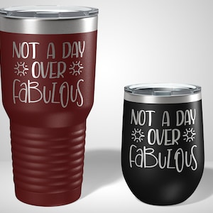 May include: Two stainless steel tumblers with a silver lid and a black and white design. The larger tumbler is maroon and the smaller tumbler is black. Both tumblers have the text "Not a day over fabulous" with a sun graphic on either side of the word "over".