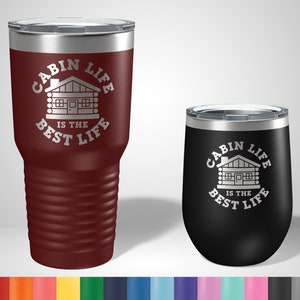 May include: Two stainless steel tumblers with a cabin life design. The larger tumbler is maroon and the smaller tumbler is black. Both tumblers have a white cabin graphic and the text "Cabin Life is the Best Life".