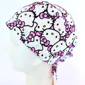 May include: A white scrub cap featuring an all-over repeating pattern of Hello Kitty characters. The design includes the iconic white cat faces with black outlines, yellow noses, and pink bows, set against a clean white background fabric.