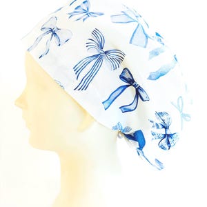 May include: A white scrub cap featuring a repeating pattern of various blue bows. The design includes bows in different shades of blue, some with solid colours and others with striped or textured details, distributed across the fabric surface.