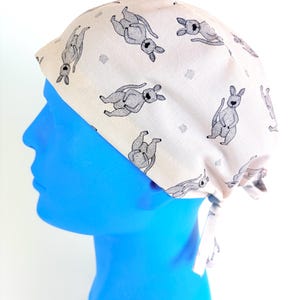 Peut inclure: Une charlotte chirurgicale beige clair ornée d'un motif répétitif de kangourous dessinés au trait noir et blanc. Le bonnet présente une calotte arrondie et des liens en tissu à l'arrière pour un ajustement réglable, avec des détails précis.